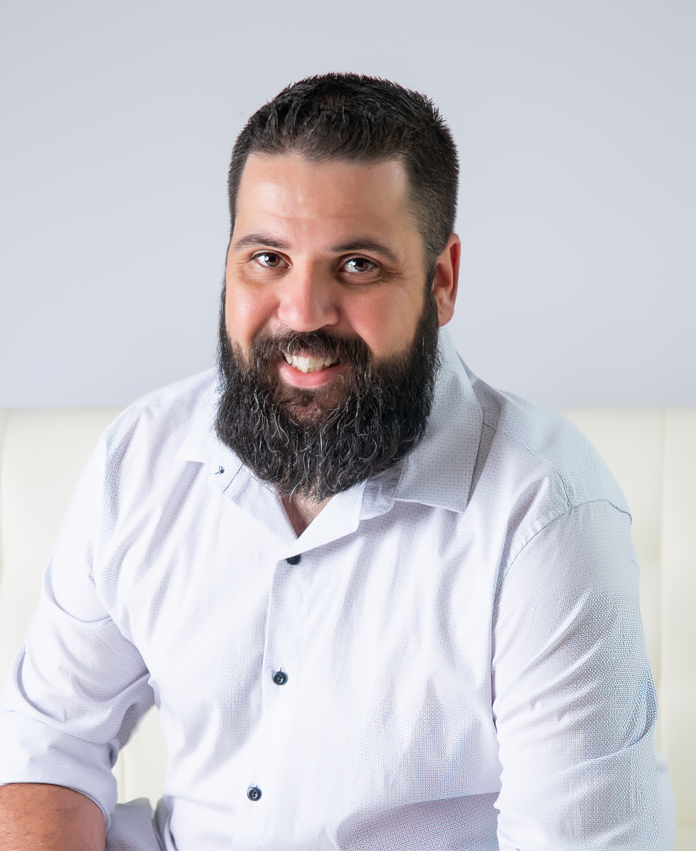 Smiling man with a beard wearing a light-colored shirt, seated against a neutral background.