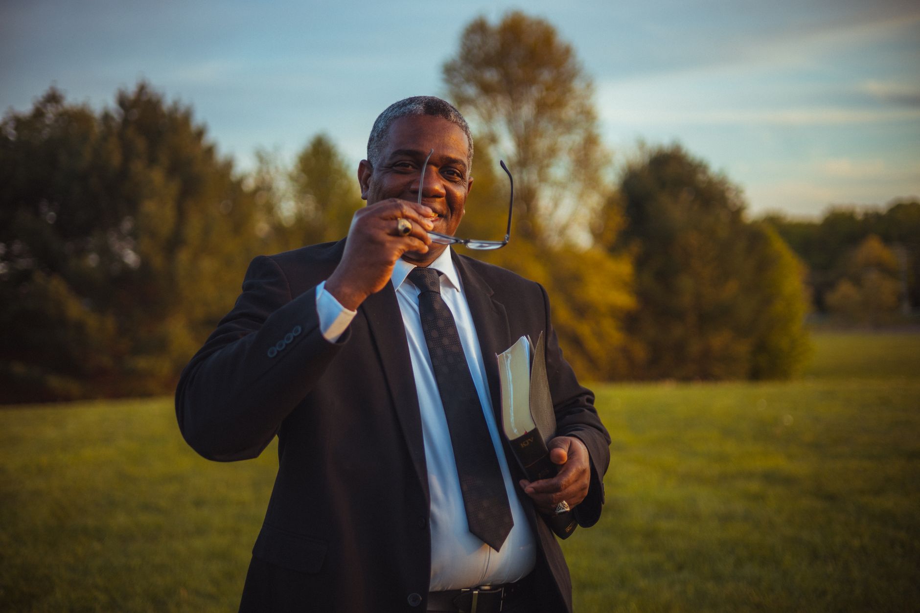 a man in black suit smiling while holding a book and eyeglasses